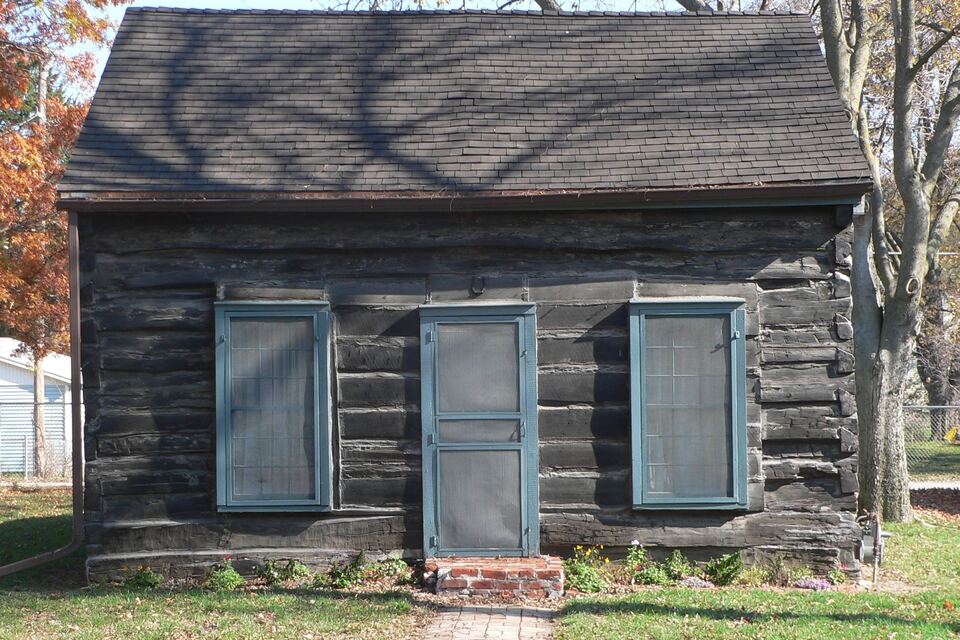 Historic weathered log cabin with blue-gray shutters on a lawn, with fall-colored trees.