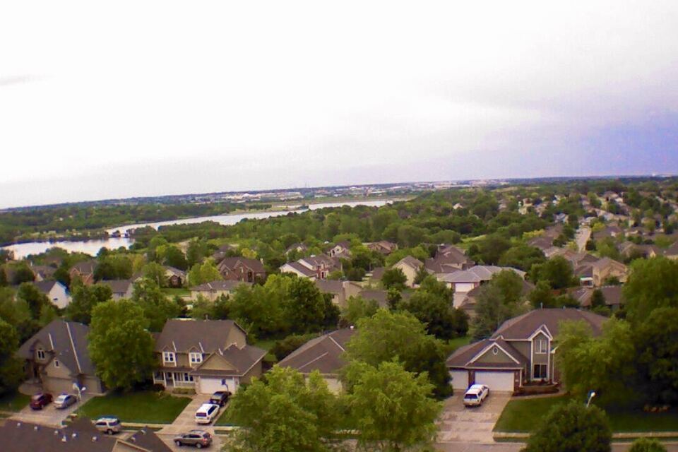 Aerial view over tree-filled suburban streets and houses in West Omaha with a lake in the distance.