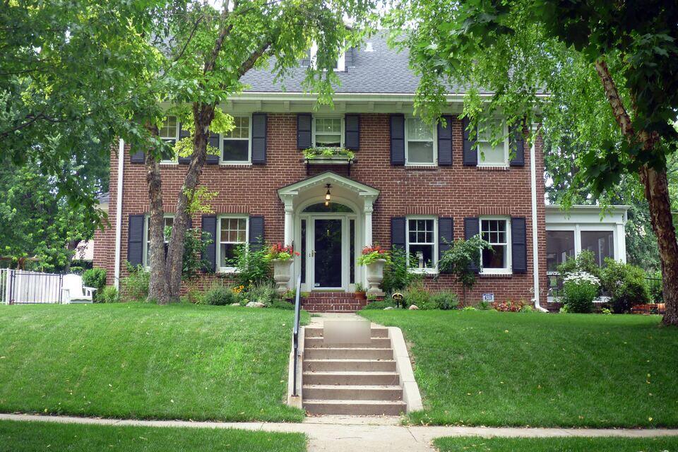 Two-story red brick colonial house with black shutters, a white front portico and concrete steps rising through a sloped green lawn, framed by mature trees.