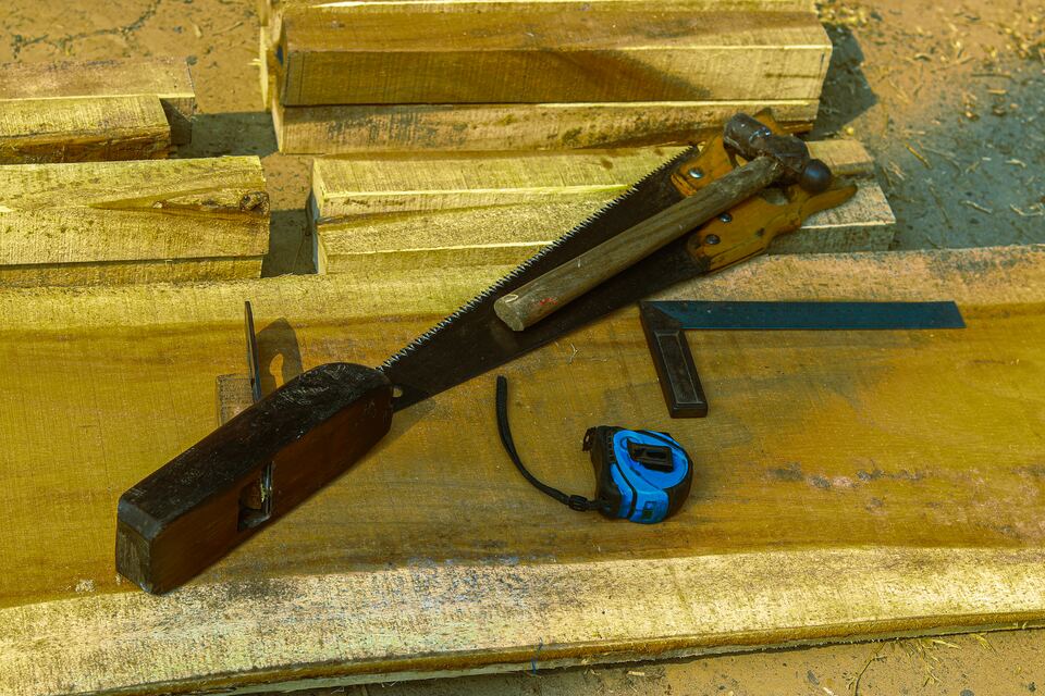 A hand saw, wooden jack plane, hammer and tape measure resting on stacked wooden planks.