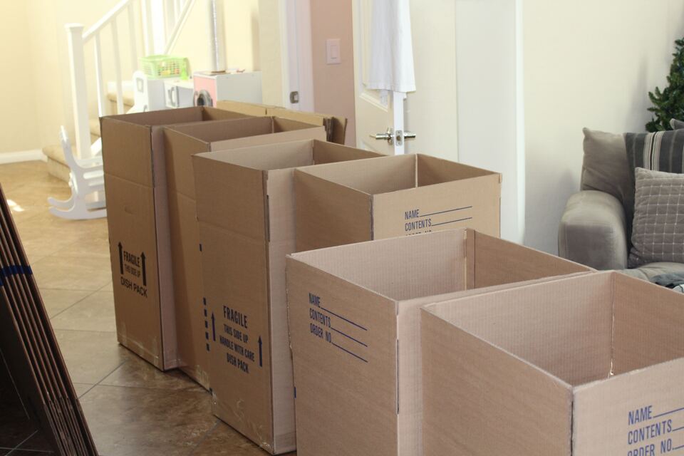 Several open and stacked cardboard moving boxes on a hardwood floor inside a home.